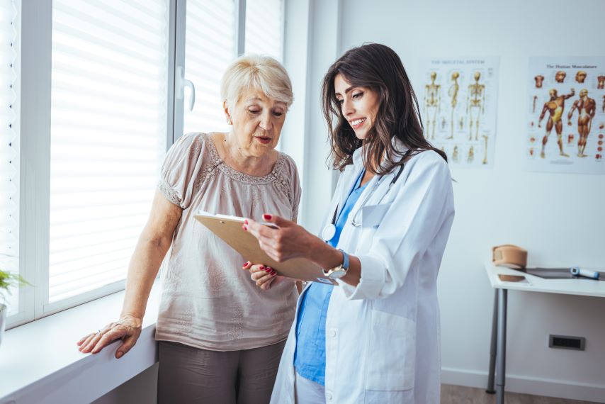 Eine Ärztin erklärt einer älteren Patientin Daten auf einem Klemmbrett.