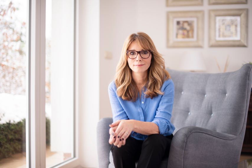 Eine Frau mit Brille und blauer Bluse sitzt auf einem grauen Sessel in einem hellen Raum